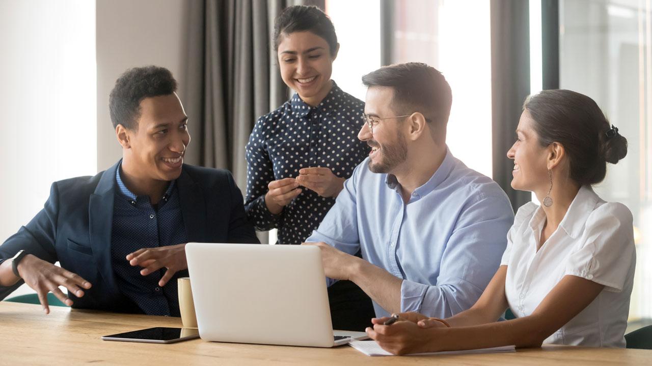 A diverse group of people sits around a conference table looking at a laptop and talking